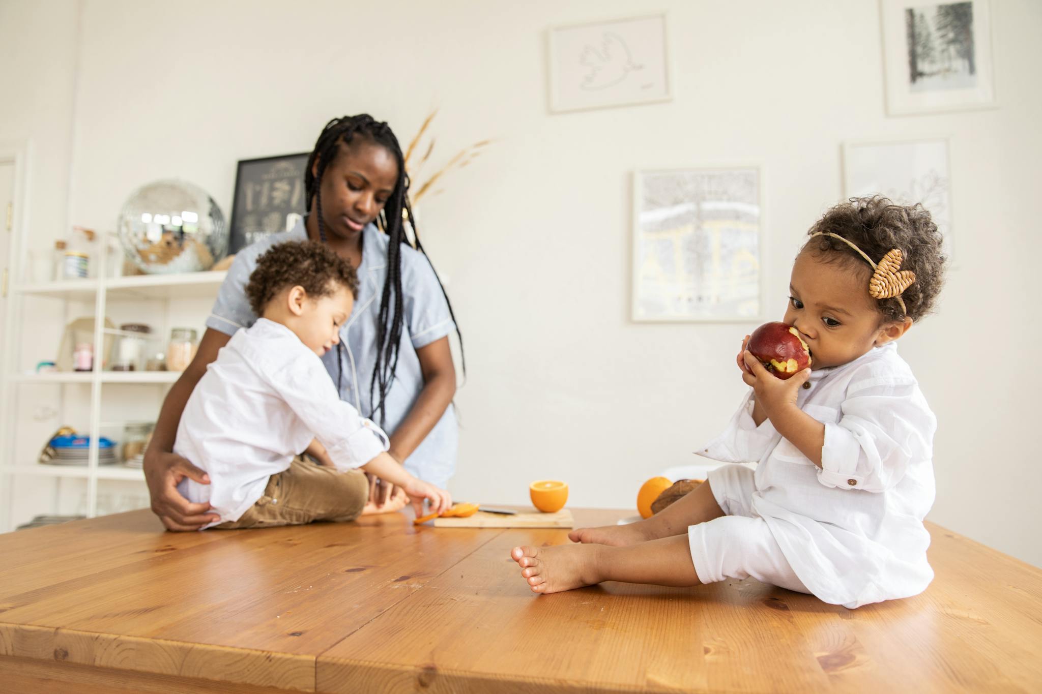 A mother with her young children at home enjoying apples and oranges on a wooden table.